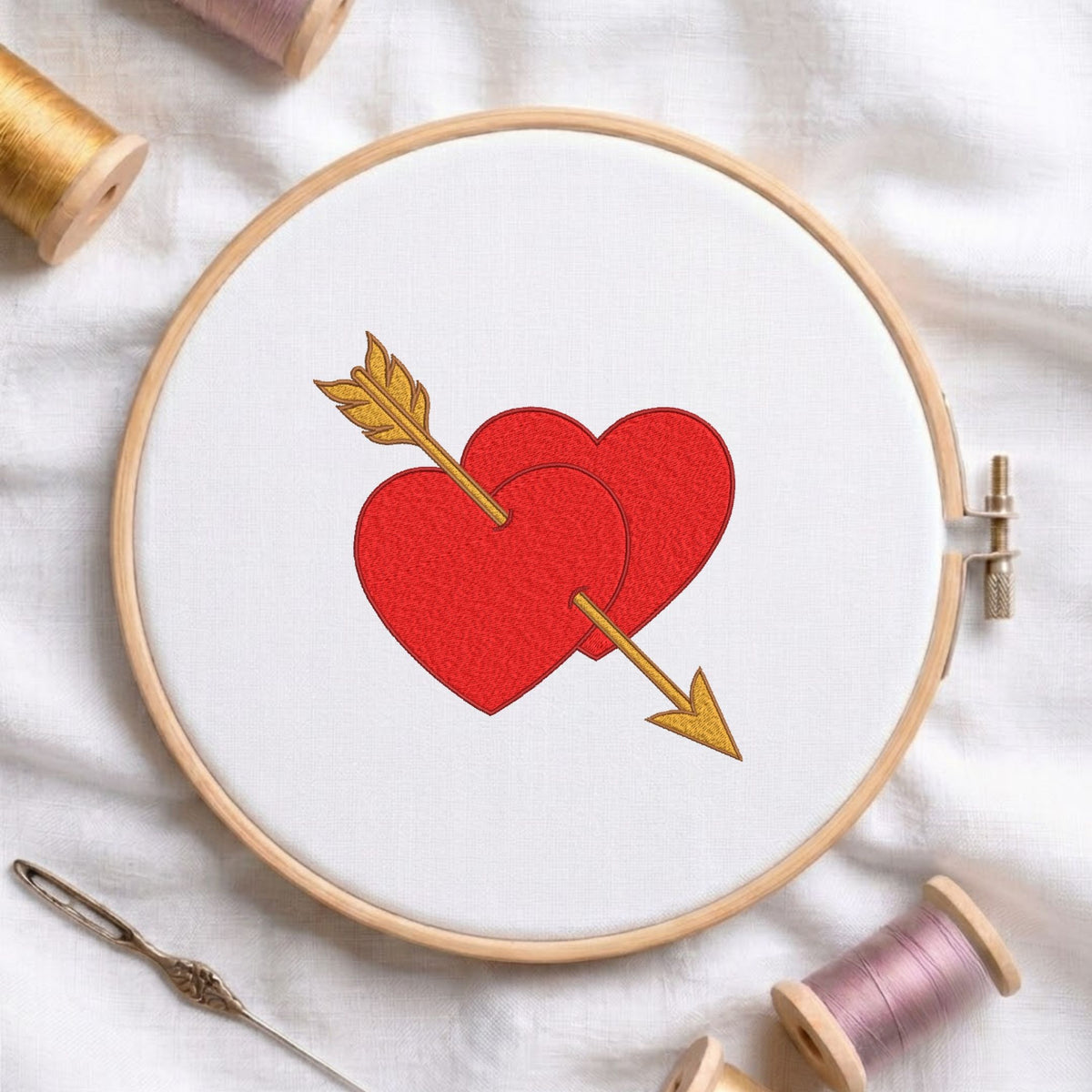 A wooden embroidery hoop holding white fabric with a red embroidered heart design pierced by a gold arrow. Spools of thread, a needle, and soft white fabric are arranged around the hoop.