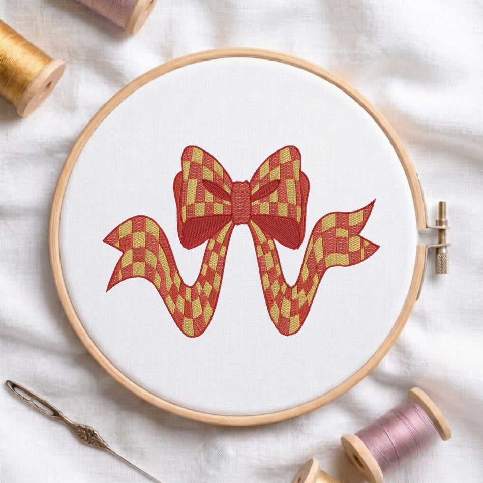 A circular embroidery hoop holding white fabric with a stitched red and gold checkered bow design, surrounded by spools of thread and a needle on a white fabric background.