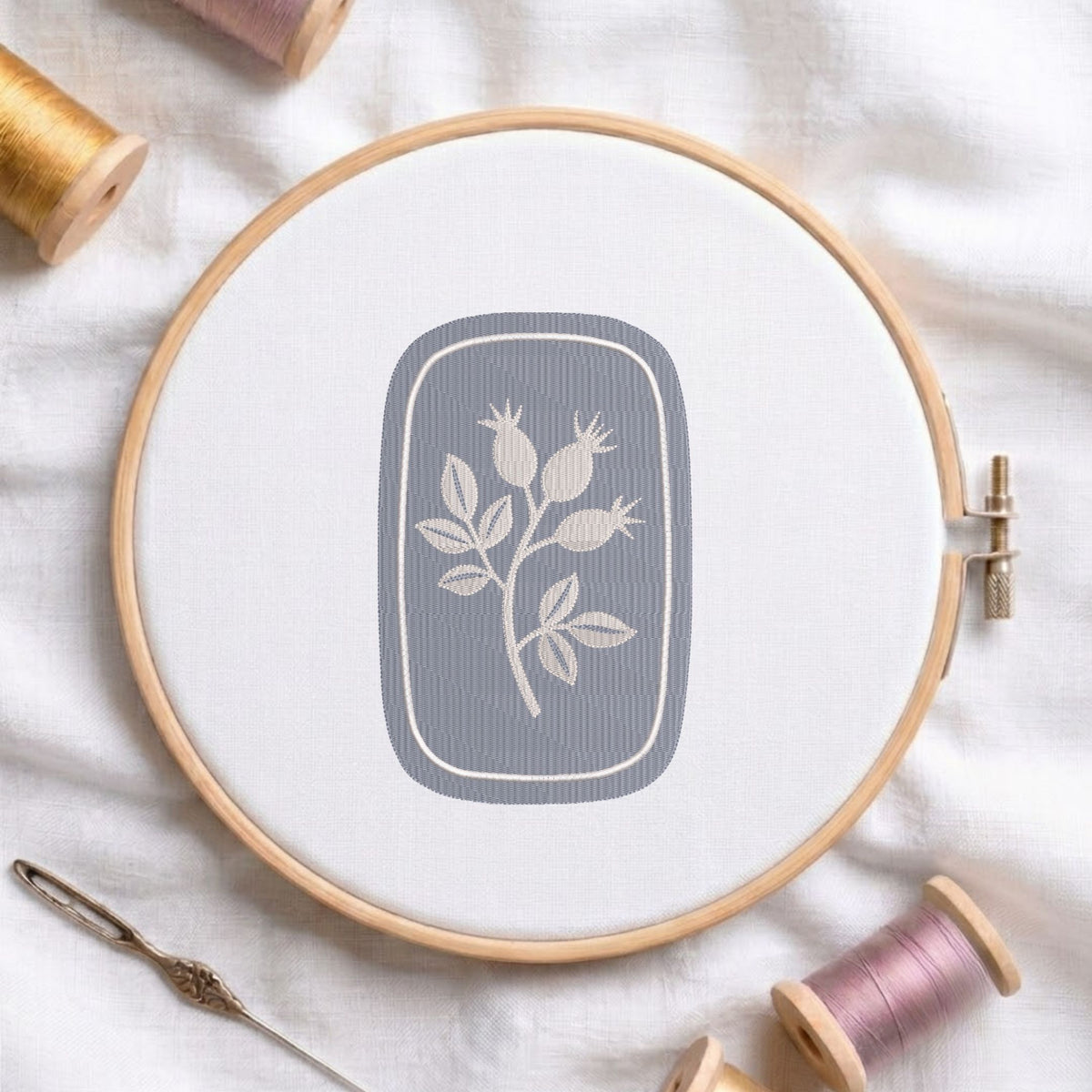 Vintage botanical rosehip branch machine embroidery design framed inside a rounded rectangular border, displayed in a wooden hoop on white fabric with thread spools and a needle