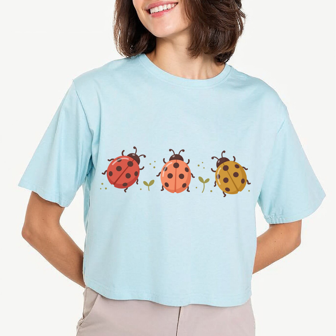 A woman wearing a light blue short-sleeve crop top t-shirt with a graphic of three ladybugs in red, orange, and yellow on the front, paired with beige pants, against a plain off-white background.