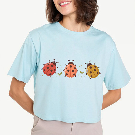 A woman wearing a light blue short-sleeve crop top t-shirt with a graphic of three ladybugs in red, orange, and yellow on the front, paired with beige pants, against a plain off-white background.