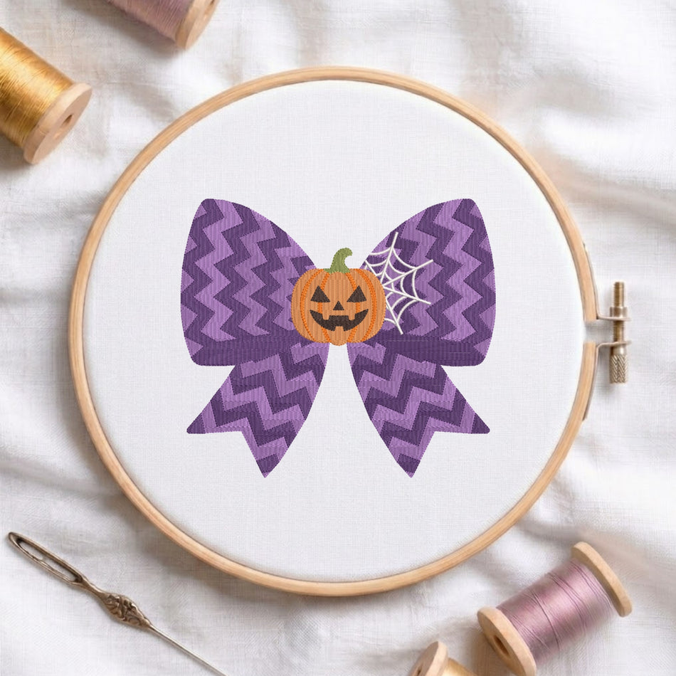 white fabric stretched in a wooden embroidery hoop, featuring an embroidered design of a purple chevron-patterned bow centered with a smiling jack-o'-lantern pumpkin and a white spiderweb.