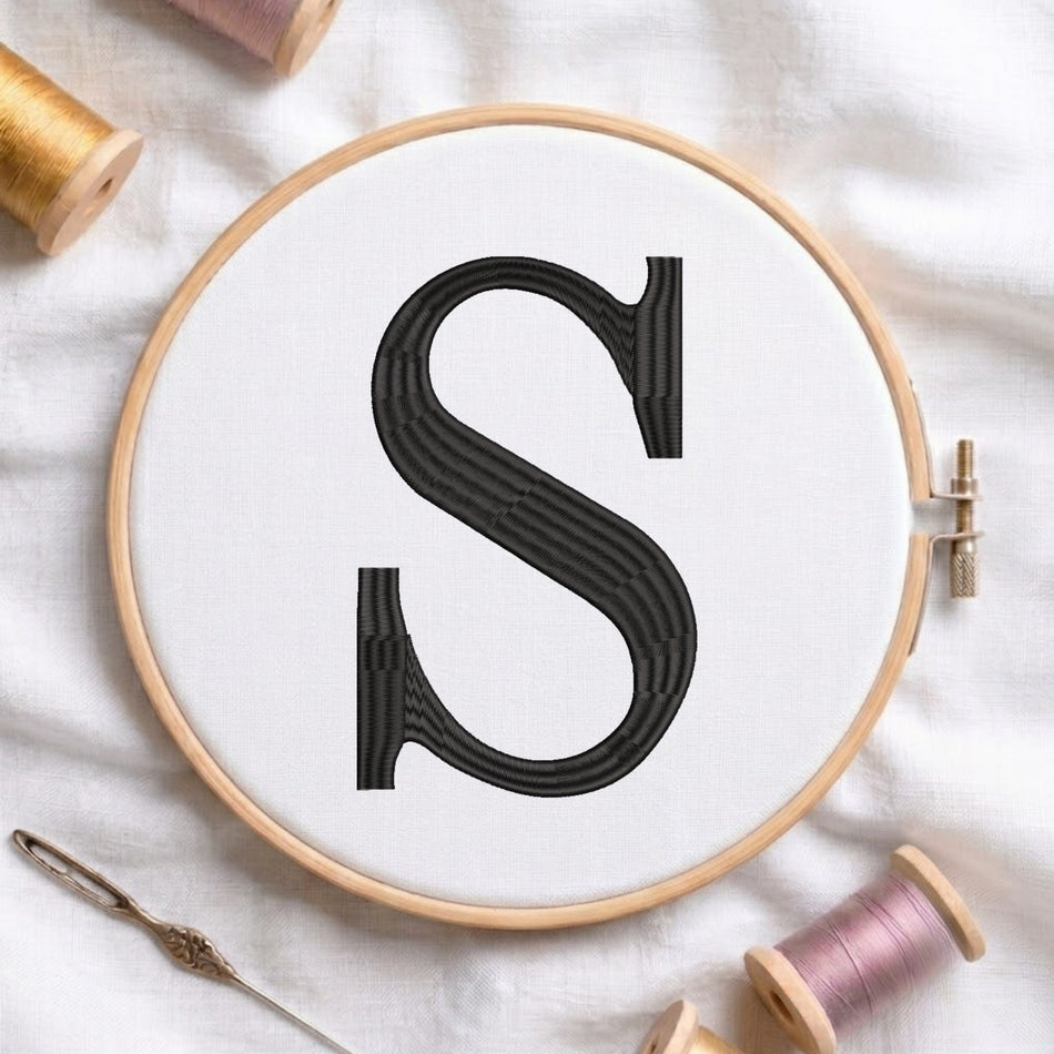 A wooden embroidery hoop holding white fabric with a large black embroidered letter S, styled on a white cloth with embroidery thread spools and a needle around it.