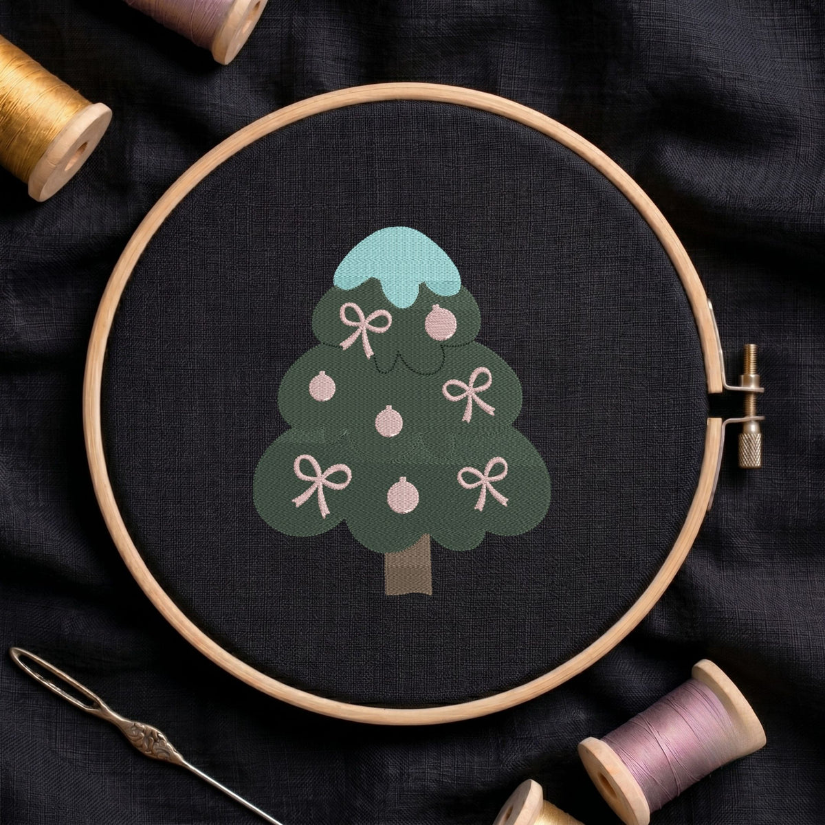 A wooden embroidery hoop holds a dark fabric with a stylized Christmas tree design in muted green, light blue, pink, and brown. Spools of thread and a needle are arranged around it on dark fabric.