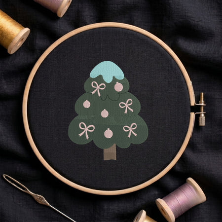 A wooden embroidery hoop holds a dark fabric with a stylized Christmas tree design in muted green, light blue, pink, and brown. Spools of thread and a needle are arranged around it on dark fabric.