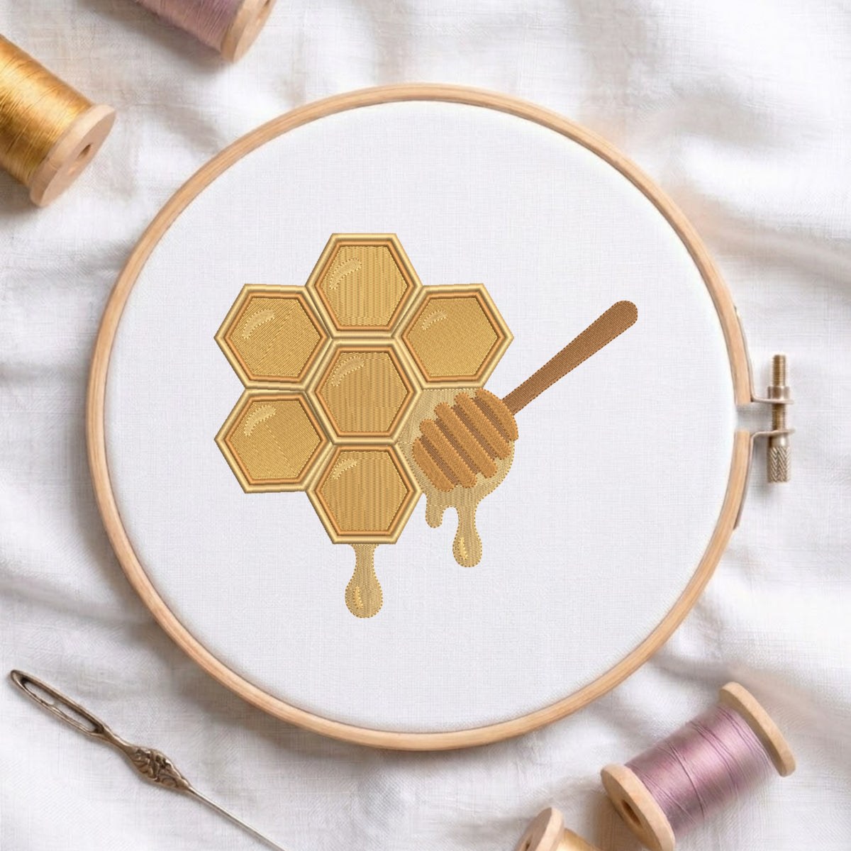 A circular embroidery hoop holds white fabric with a stitched honeycomb and dripping honey design in warm gold and tan tones. A wooden honey dipper is stitched over the center.