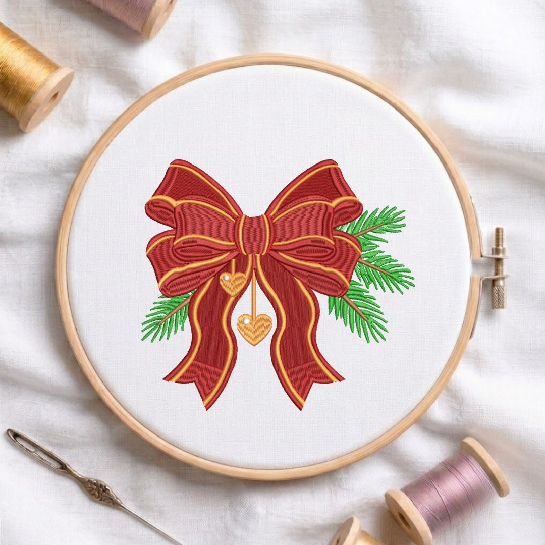 A circular embroidery hoop on white fabric displays a festive red bow with green pine branches and gold hanging hearts. Spools of thread and a needle are arranged around the hoop.