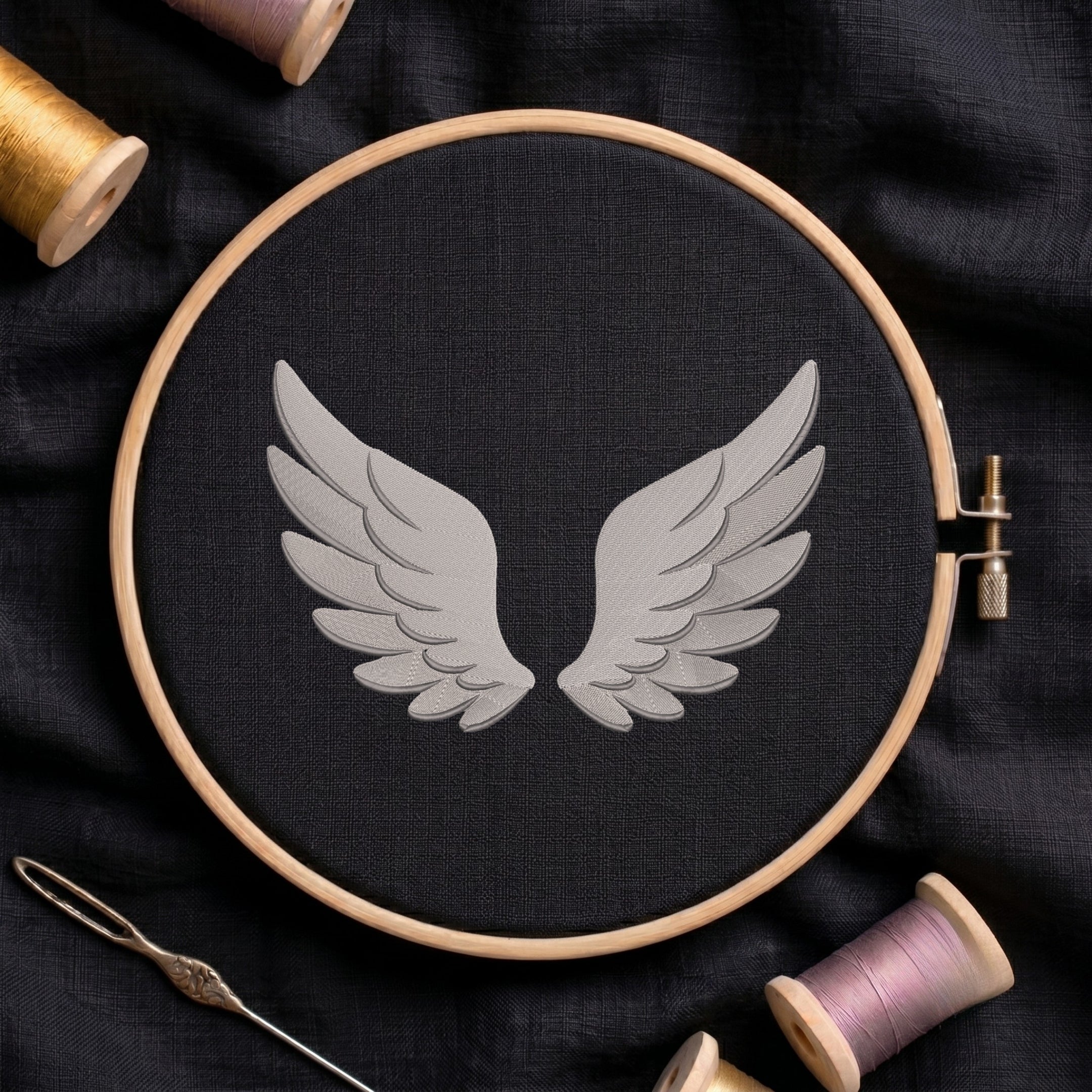 A circular embroidery hoop holding black fabric with a gray wing motif in the center, surrounded by spools of thread and a needle on a dark fabric background.