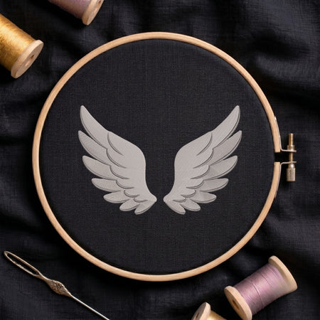 A circular embroidery hoop holding black fabric with a gray wing motif in the center, surrounded by spools of thread and a needle on a dark fabric background.