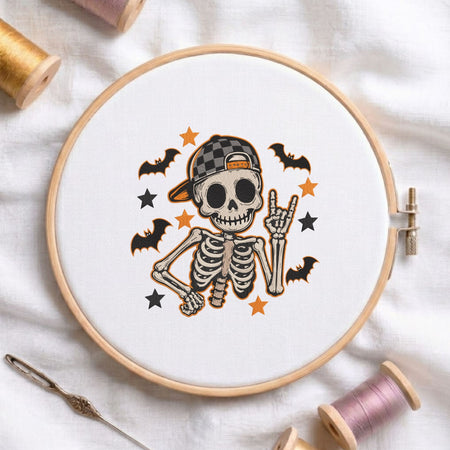 Cute skeleton wearing a cap and making a rock sign with bats and stars machine embroidery design shown in a wooden embroidery hoop on white fabric with thread spools and a needle