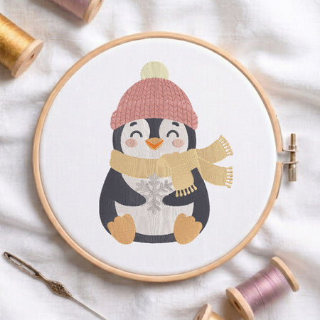 A circular embroidery hoop holds a white fabric with a stitched penguin wearing a pink knit hat, cream pom-pom, black-and-white body, and a beige scarf. The hoop is photographed on white fabric with spools of thread and a needle around it.