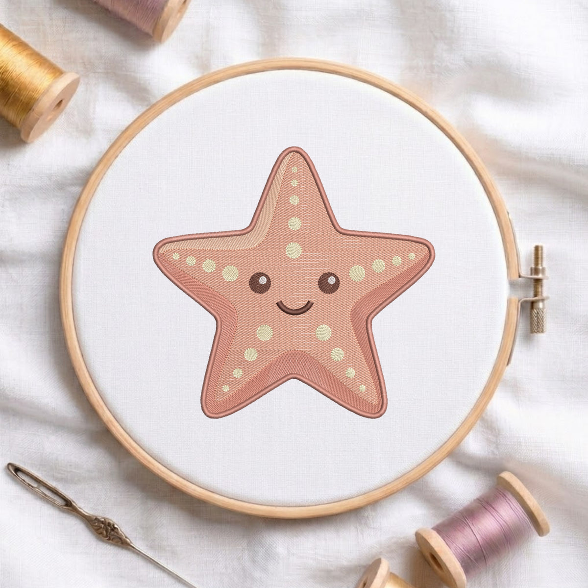 A wooden embroidery hoop holding white fabric with a cute starfish design stitched in dusty pink and cream. The hoop is styled on a white fabric background with spools of thread and a needle.