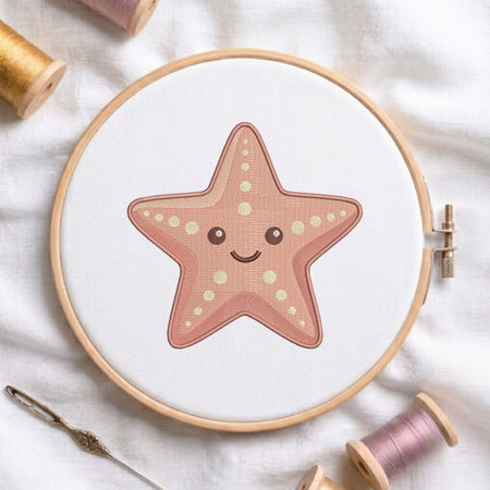A wooden embroidery hoop holding white fabric with a cute starfish design stitched in dusty pink and cream. The hoop is styled on a white fabric background with spools of thread and a needle.