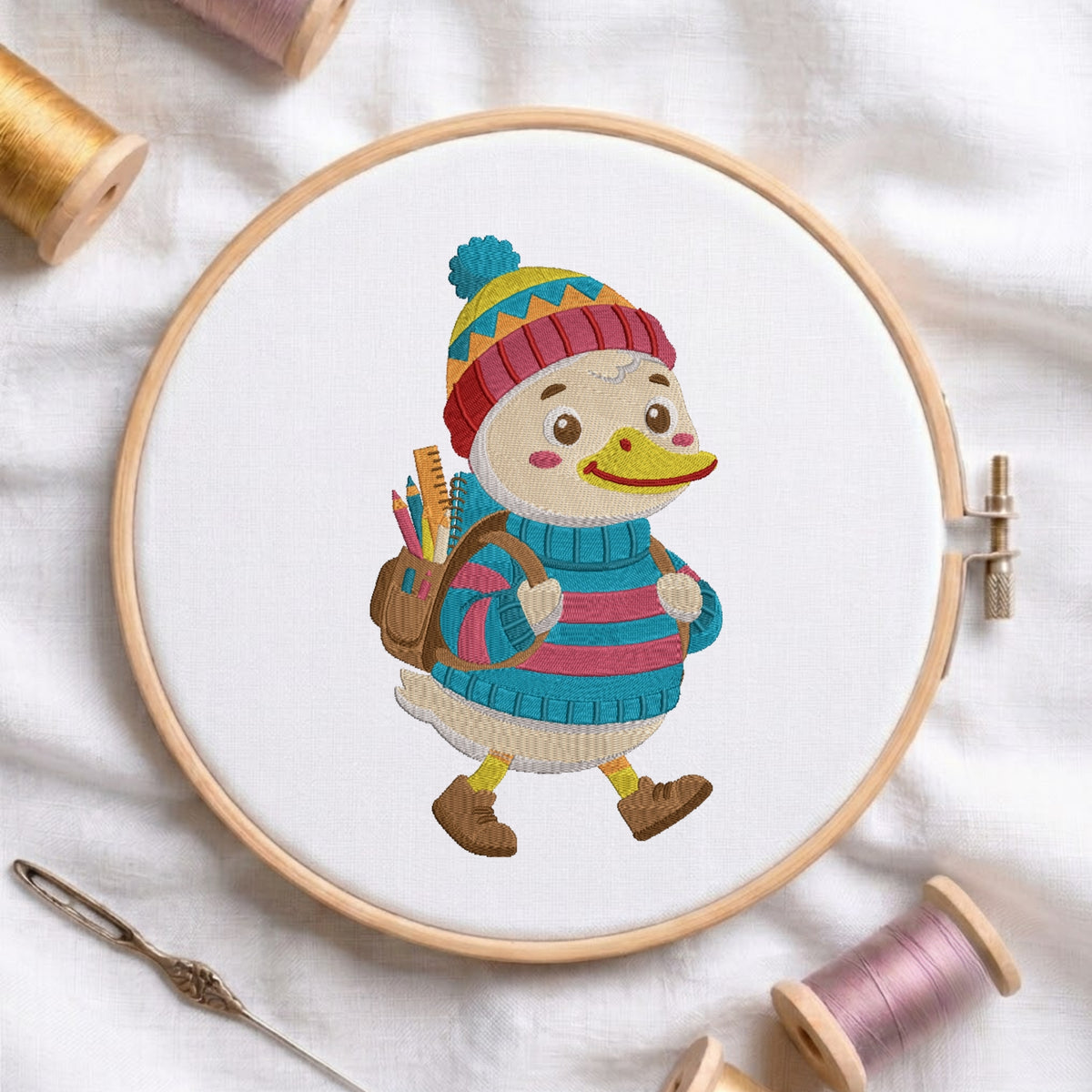 A circular embroidery hoop holds a white fabric with a colorful embroidered duck character wearing a striped knit hat and blue sweater, surrounded by spools of thread and a needle on a white fabric background.