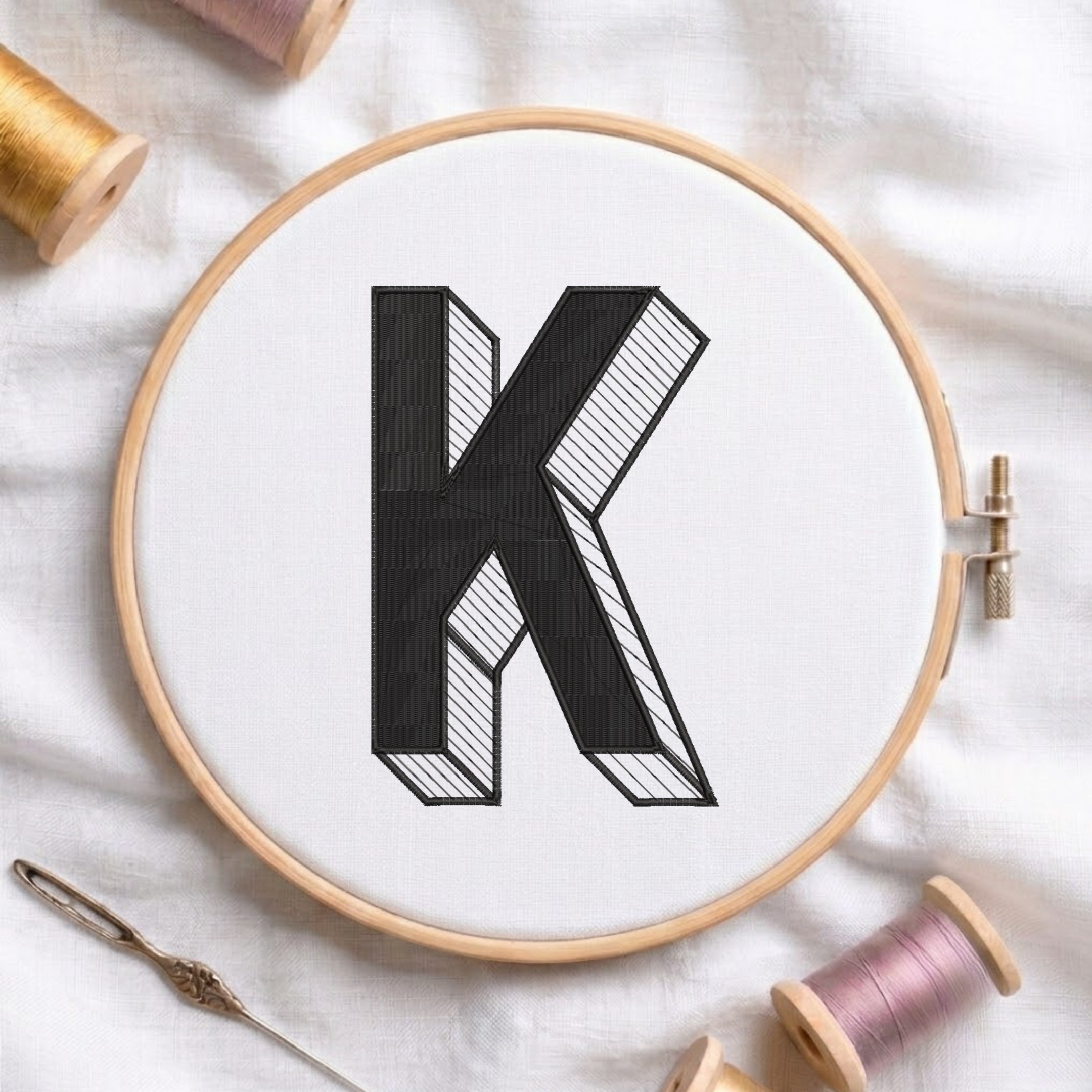 A wooden embroidery hoop holding white fabric with a large black embroidered letter K, styled on a white cloth with spools of thread and a needle around it.