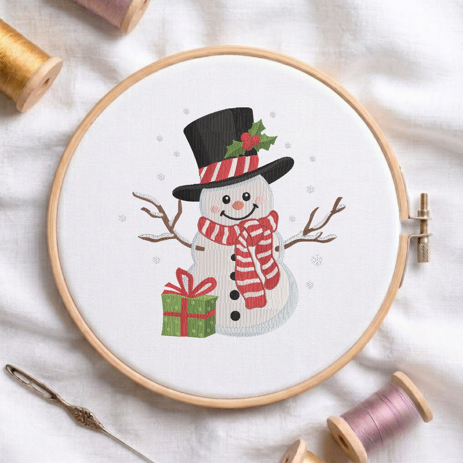 A white fabric stretched in a wooden embroidery hoop, featuring an embroidered snowman wearing a top hat with holly leaves, a striped red and white scarf, twig arms, and holding a green gift box with a red ribbon.