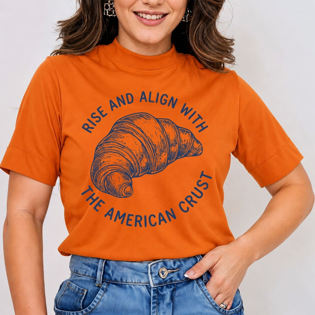 Woman wearing a burnt orange mock neck t-shirt featuring a blue retro line art graphic of a croissant with the circular text "Rise and Align with the American Crust", tucked into blue jeans.
