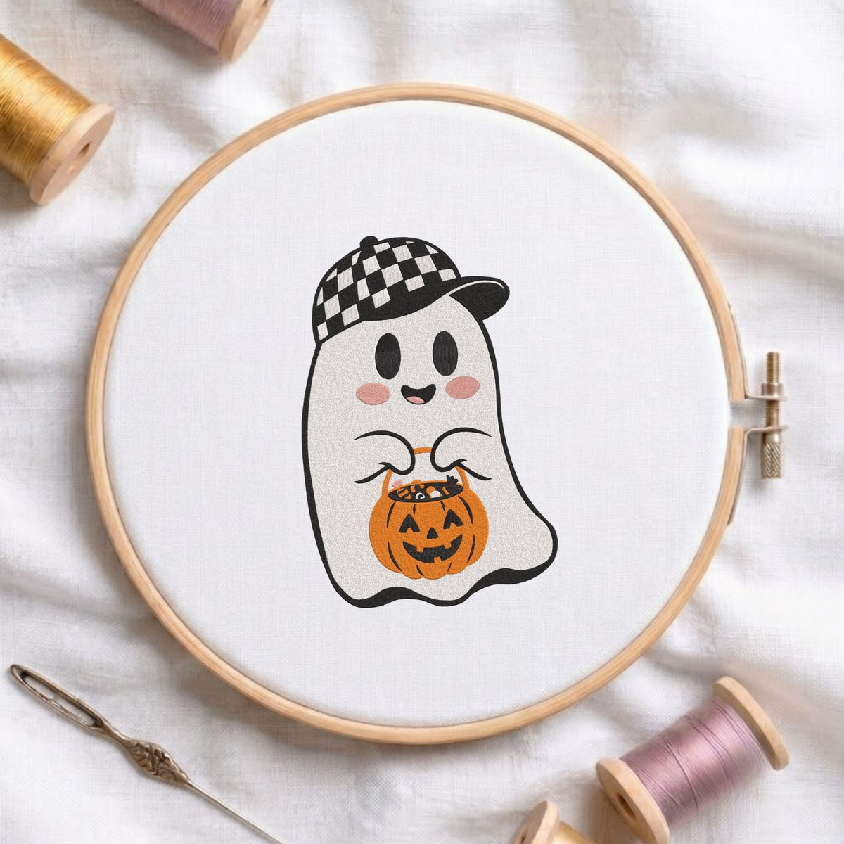 Cute ghost wearing a black and white checkered baseball cap, holding a jack-o-lantern pumpkin candy pail machine embroidery design shown in a wooden hoop on white fabric with thread spools and a needle