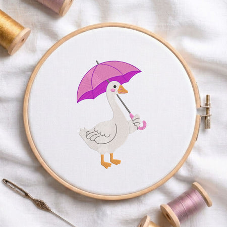 t: Cute duck holding a purple umbrella machine embroidery design displayed in a wooden hoop on white fabric with thread spools and a needle.