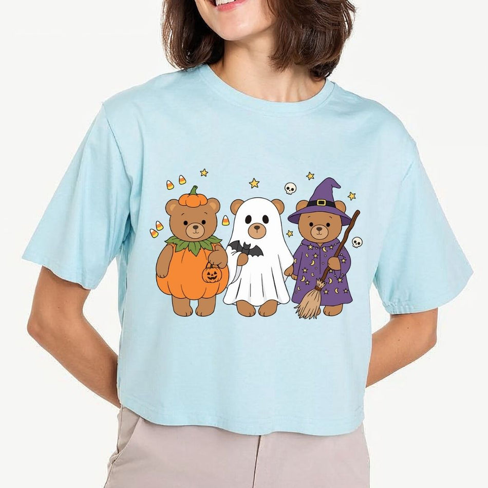 A woman wearing a light blue short-sleeve t-shirt with a graphic of three teddy bears in Halloween costumes (pumpkin, ghost, and witch). The background is a clean, off-white studio setting.