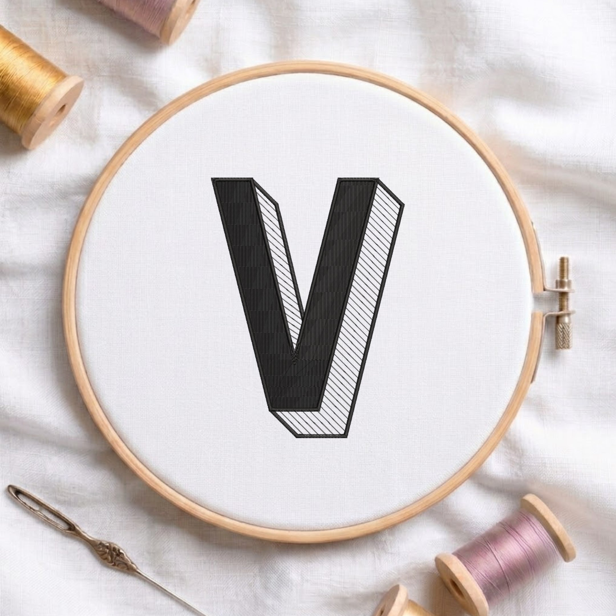 A wooden embroidery hoop holding white fabric with a large black letter V, surrounded by spools of thread and a needle on a white fabric background.