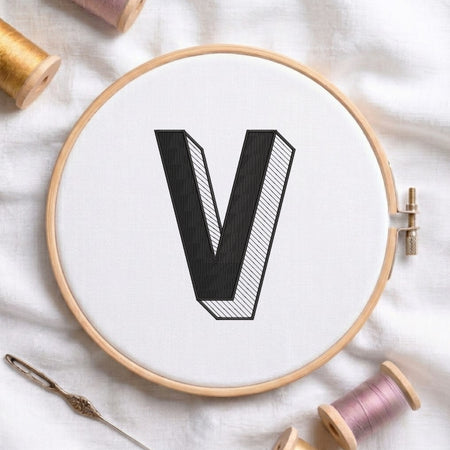 A wooden embroidery hoop holding white fabric with a large black letter V, surrounded by spools of thread and a needle on a white fabric background.