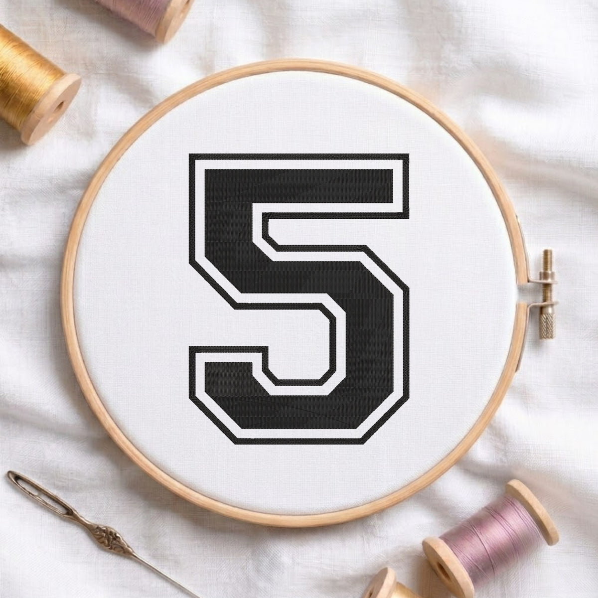 A wooden embroidery hoop holding white fabric with a large black number 5 outlined in white, styled on a white fabric surface with embroidery thread spools and a needle around it.