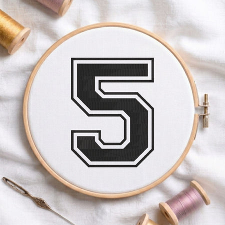 A wooden embroidery hoop holding white fabric with a large black number 5 outlined in white, styled on a white fabric surface with embroidery thread spools and a needle around it.