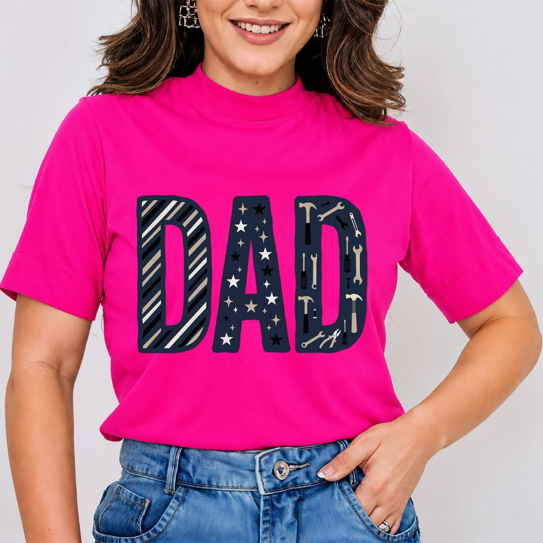 A woman wearing a bright pink mock-neck t-shirt with a large 'DAD' graphic across the chest in navy blue with white stars and tool patterns, paired with blue jeans.