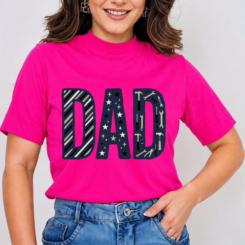 A woman wearing a bright pink mock-neck t-shirt with a large 'DAD' graphic across the chest in navy blue with white stars and tool patterns, paired with blue jeans.