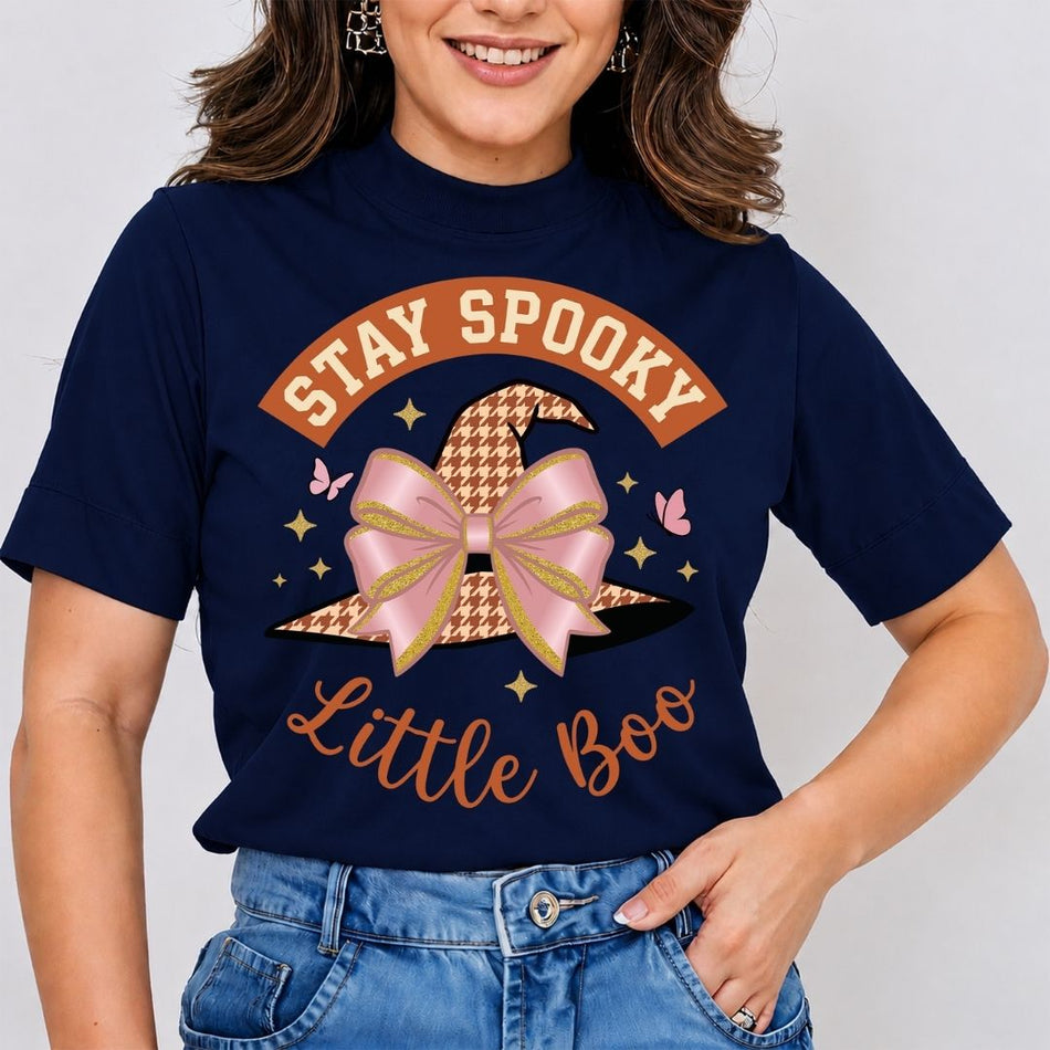 A woman wearing a navy blue graphic t-shirt with a 'Stay Spooky Little Boo' design featuring a pink and gold bow, stars, and a houndstooth pattern, paired with blue jeans.
