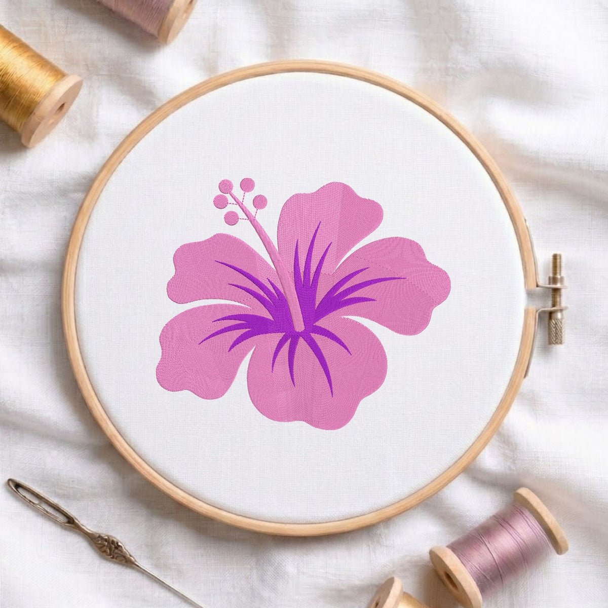 A wooden embroidery hoop holding white fabric with a large pink hibiscus flower design in the center. A metal needle lies at the lower left, and spools of thread are arranged around the hoop on a white fabric background.