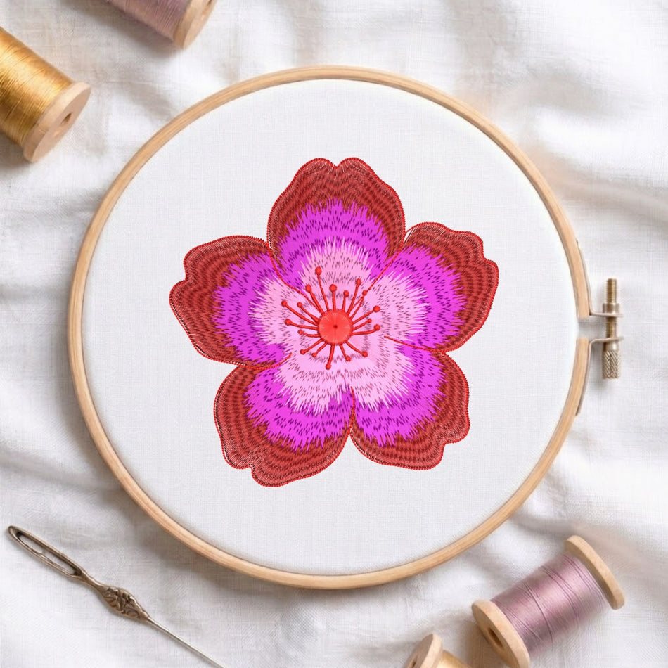 A circular embroidery hoop holding white fabric with a stylized flower design in shades of red, pink, and purple. Spools of thread, a needle, and soft fabric are arranged around it in a styled flat-lay.
