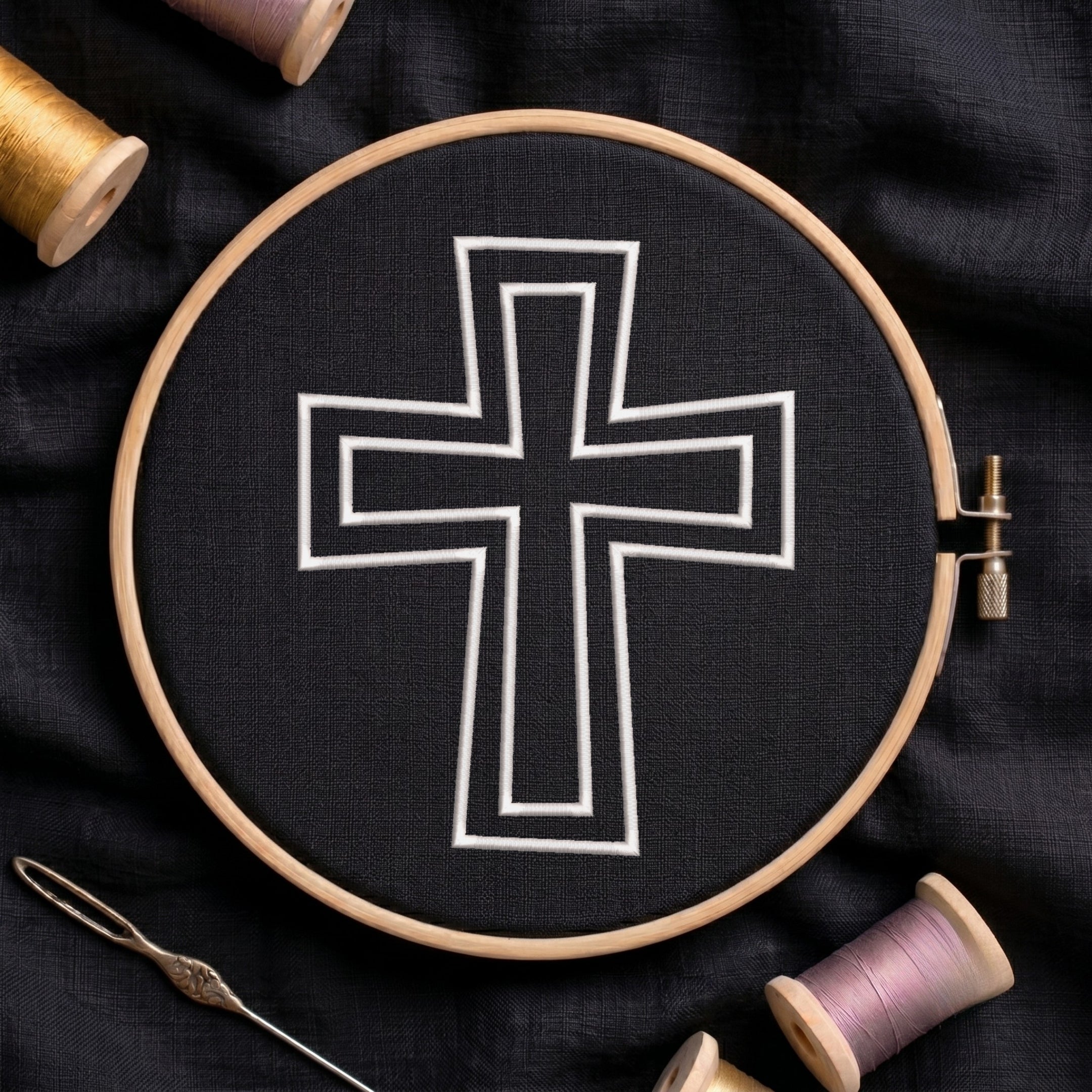 An embroidery hoop on a black cloth displays a finished embroidery design of a clean white outlined Christian cross, flanked by spools of thread and a needle.