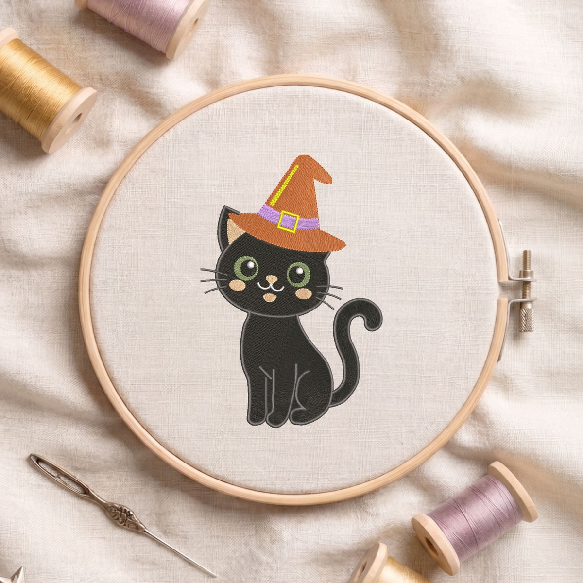 Cute black cat wearing an orange witch hat with a purple band machine embroidery design, displayed inside a round wooden hoop on white linen fabric alongside thread spools and a needle.