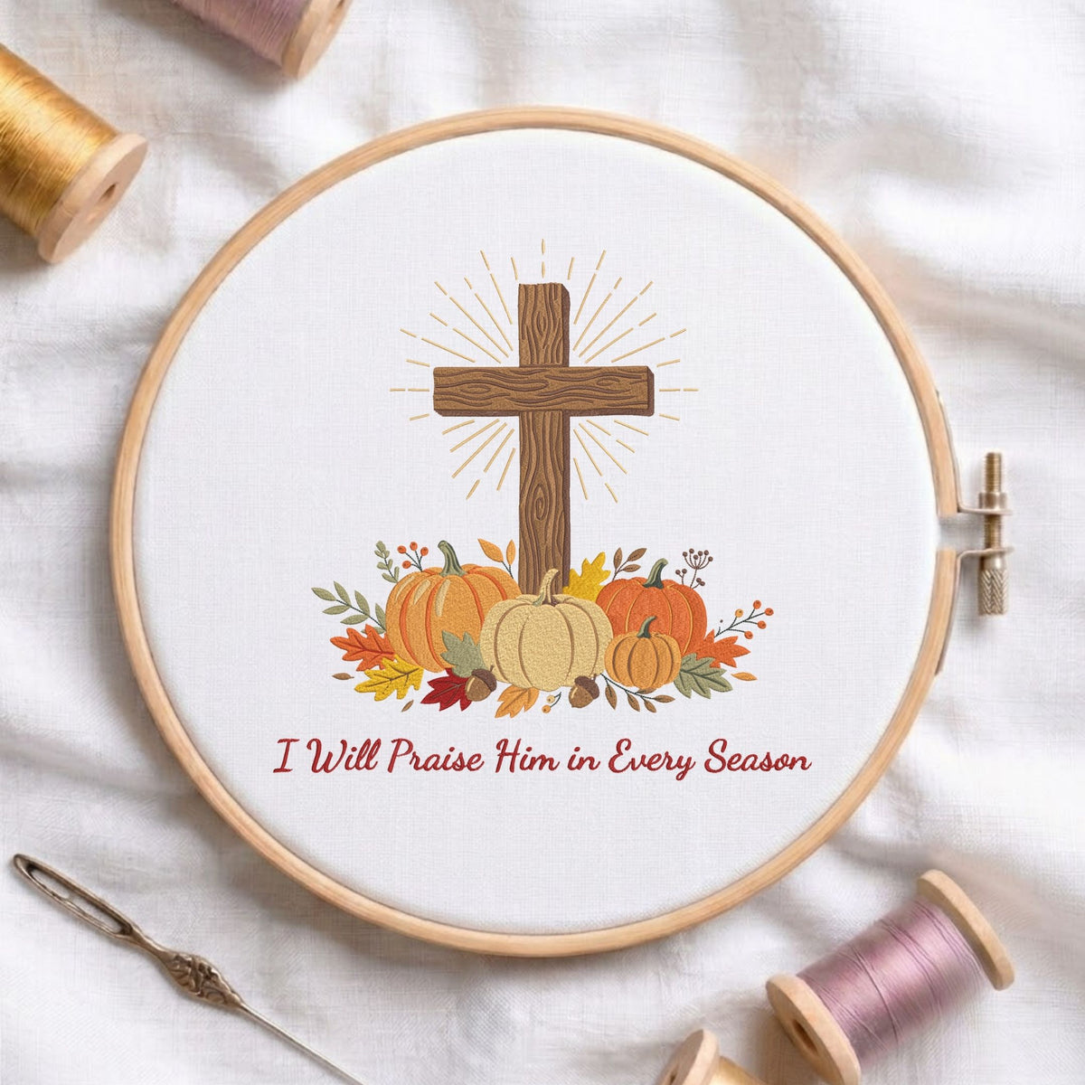 Christian cross with pumpkins, fall leaves, and the quote "I Will Praise Him in Every Season" machine embroidery design shown in a wooden embroidery hoop on white fabric with thread spools and a needle