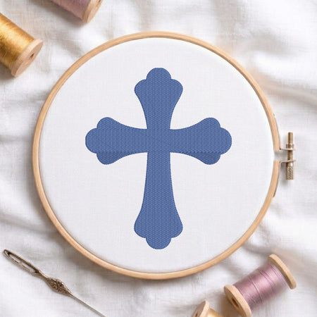 A wooden embroidery hoop holding white fabric with a blue cross design, surrounded by spools of thread and a needle on a white fabric background.