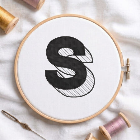 A wooden embroidery hoop holding white fabric with a large black letter S design, surrounded by spools of thread and a needle on a white fabric background.