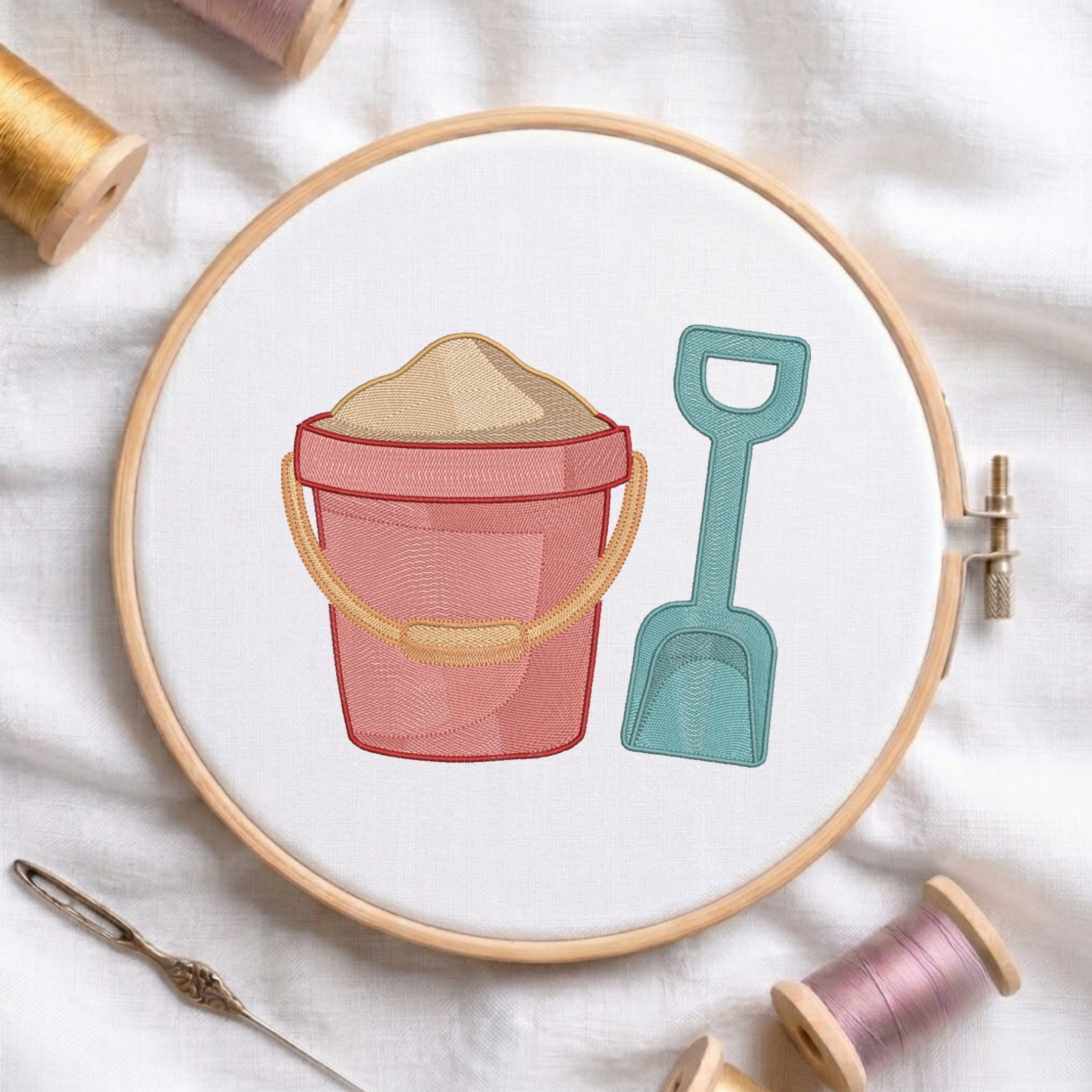 A circular embroidery hoop on white fabric displays a beach-themed design of a pink bucket filled with sand and a teal shovel. Surrounding the hoop are spools of thread, a needle, and a pin.