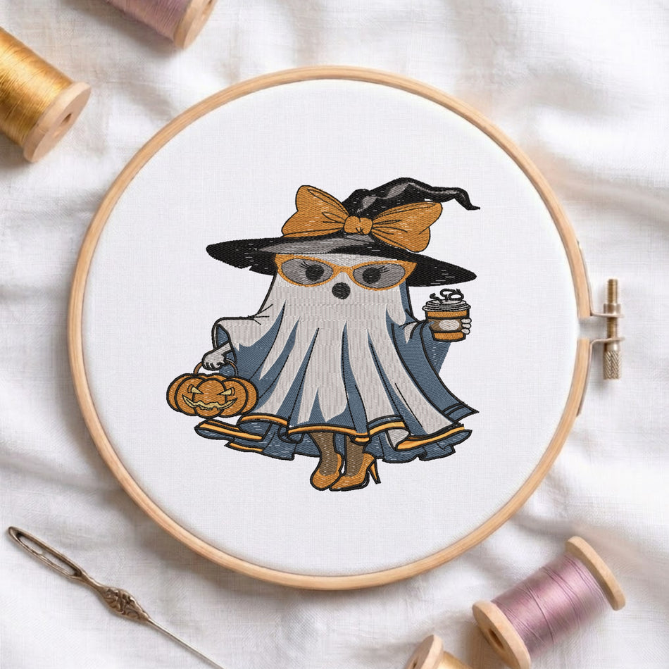 A circular embroidery hoop displays a cartoon ghost dressed as a witch with a black hat, orange bow, blue dress, and holding a pumpkin and coffee cup. The hoop is styled on a white fabric background with spools of thread and a needle.