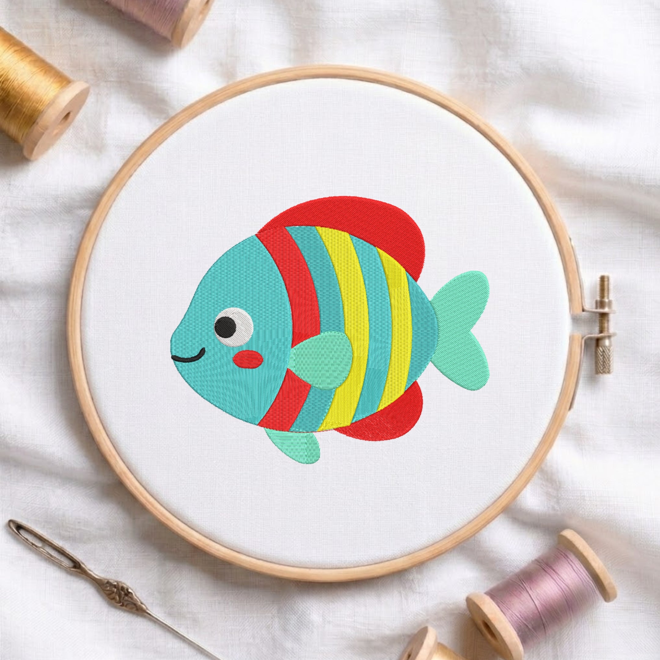 A colorful embroidered fish design inside a wooden embroidery hoop, surrounded by spools of thread and a needle on a white fabric background.