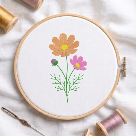 Peach and pink cosmos wildflowers with green stems embroidered inside a round wooden hoop with thread spools.