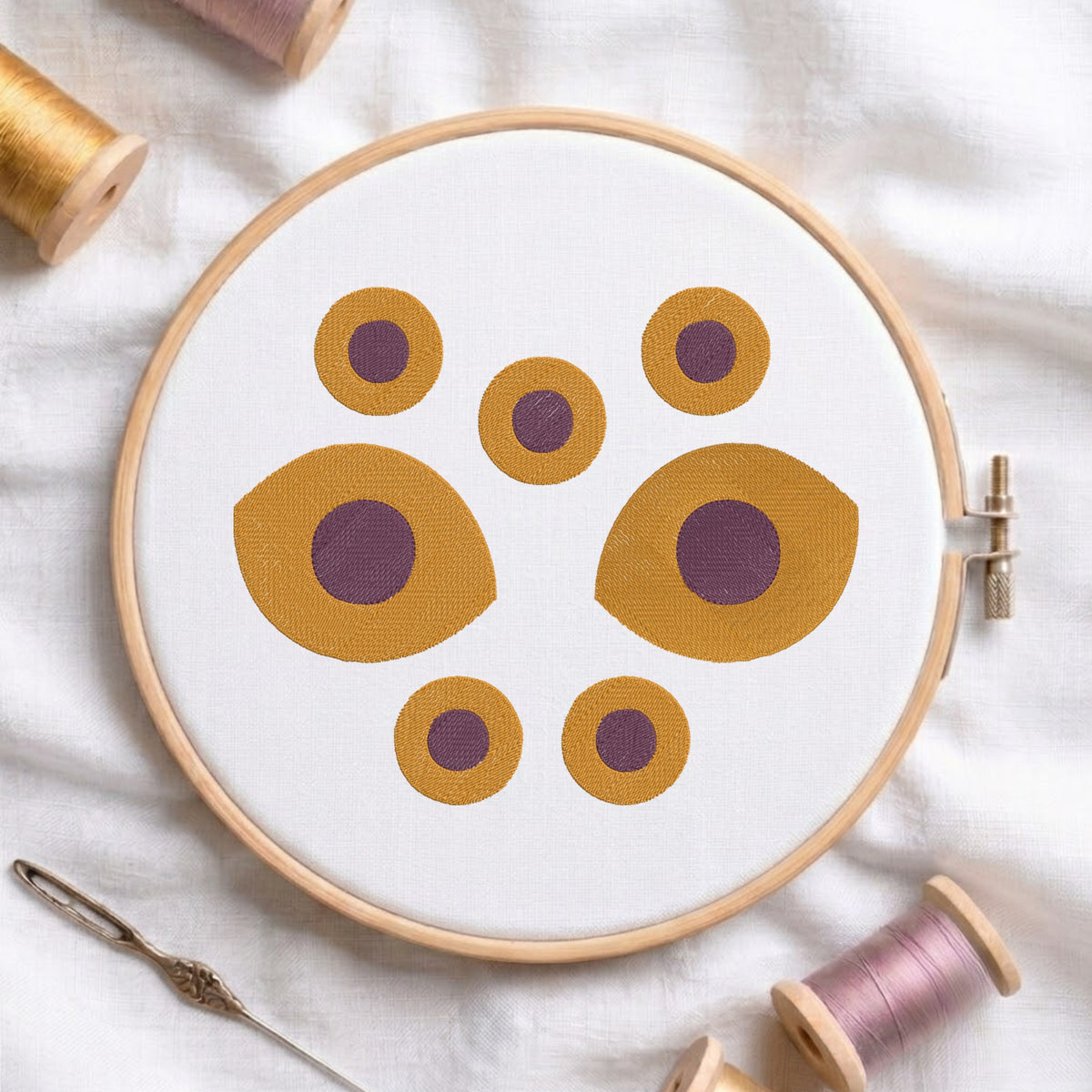 A circular embroidery hoop holding white fabric with a geometric pattern of mustard yellow and purple circles, styled on a white fabric background with thread spools and a needle.