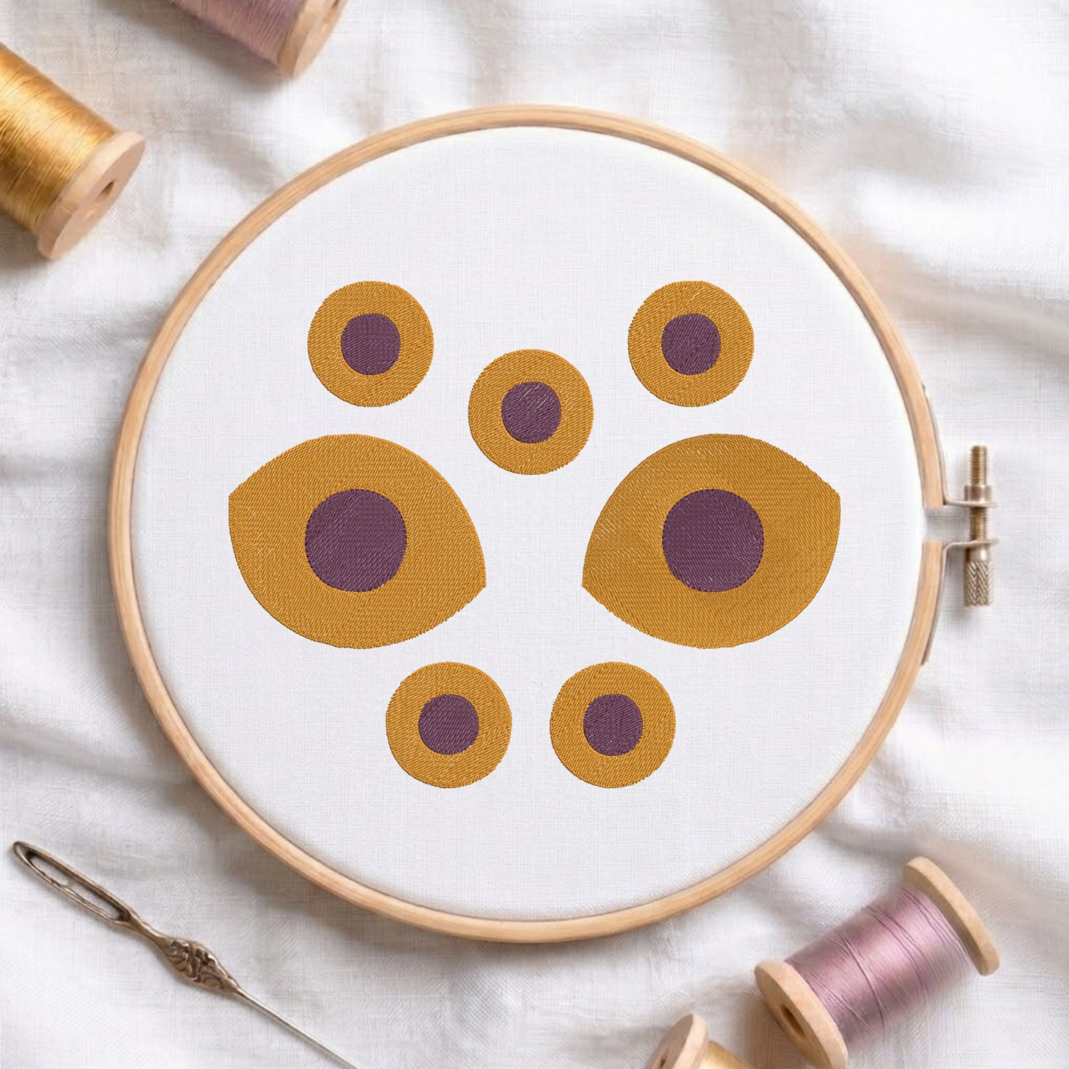 A circular embroidery hoop holding white fabric with a geometric pattern of mustard yellow and purple circles, styled on a white fabric background with thread spools and a needle.