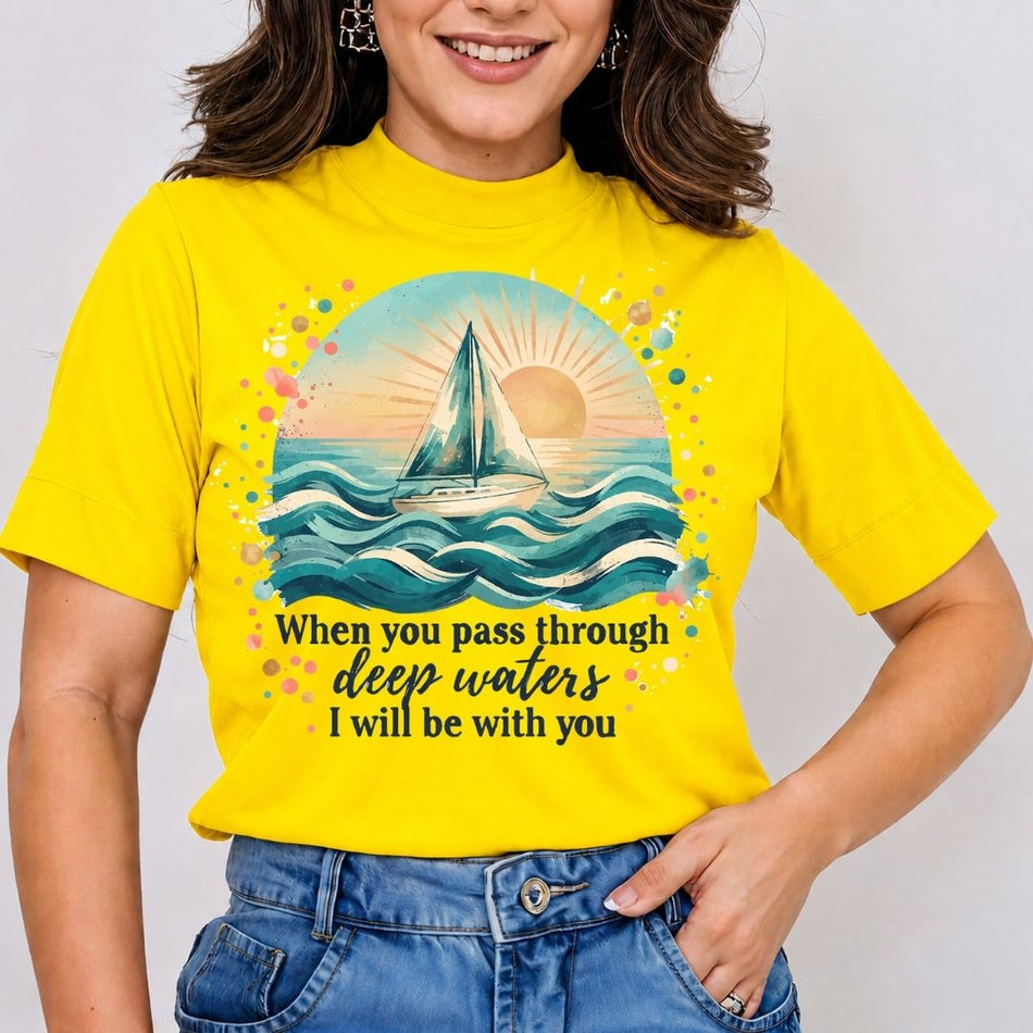 A woman wearing a bright yellow short-sleeve t-shirt with a colorful sailboat graphic and the text 'When you pass through deep waters I will be with you', paired with blue jeans against a light gray background.