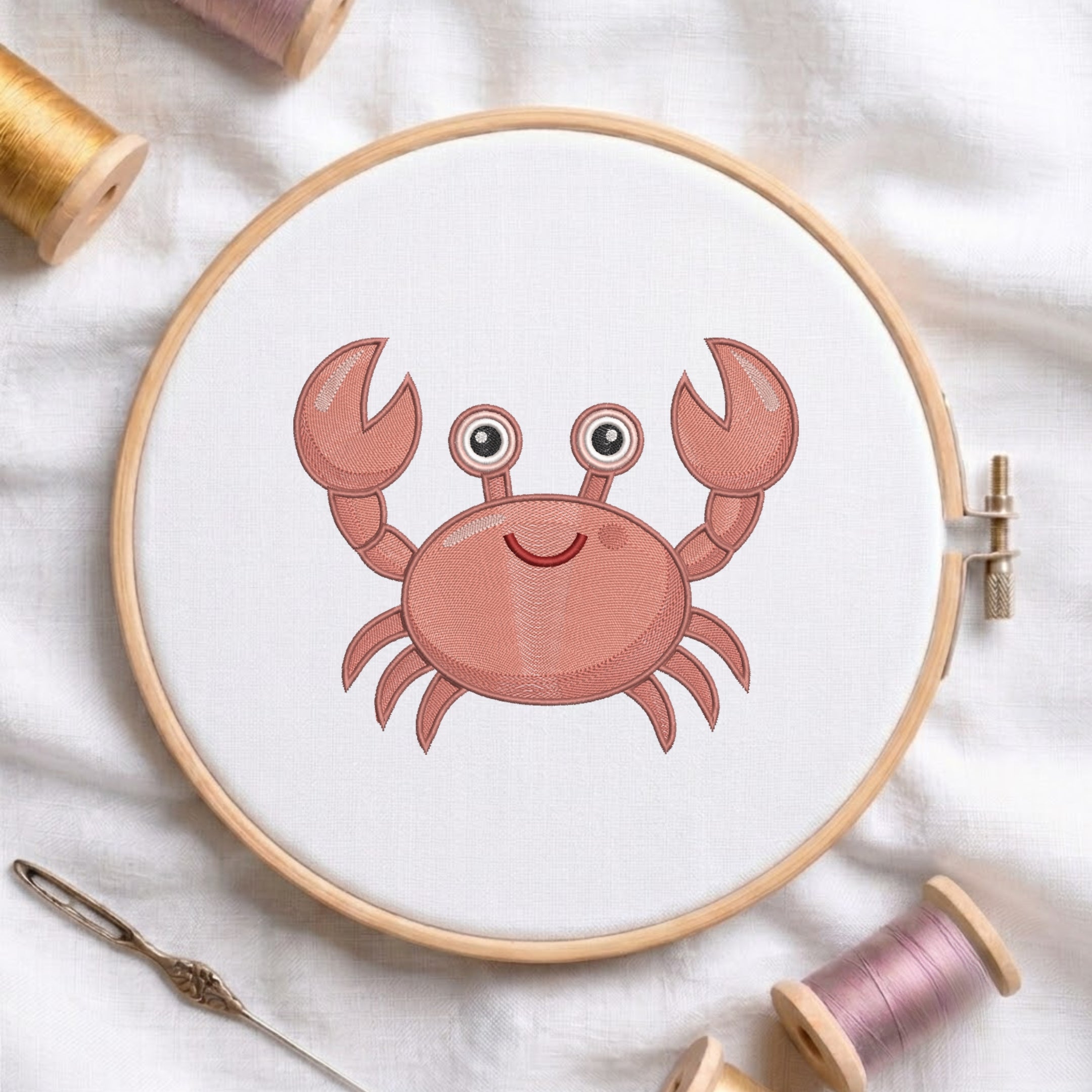 A round embroidery hoop holding white fabric with a pink cartoon crab design, surrounded by spools of thread and a needle on a white fabric background.