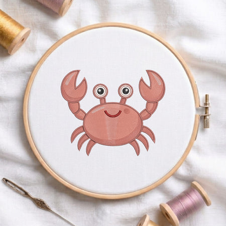 A round embroidery hoop holding white fabric with a pink cartoon crab design, surrounded by spools of thread and a needle on a white fabric background.