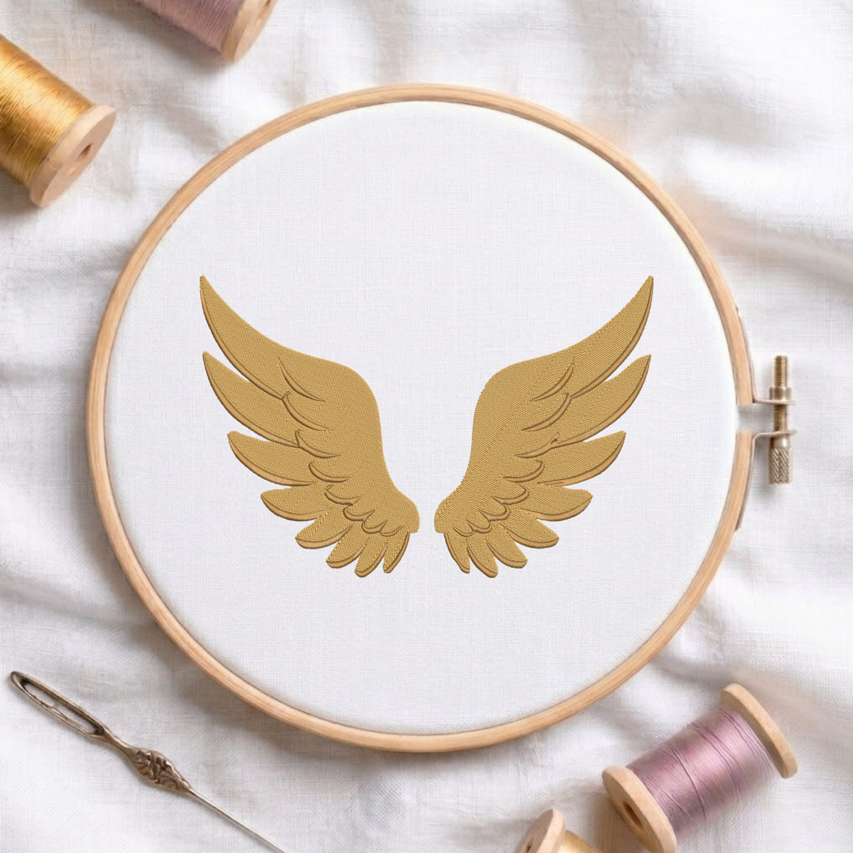 A wooden embroidery hoop holding white fabric with a gold embroidered pair of wings, styled on a white cloth with spools of thread and a needle around it.