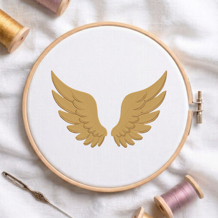 A wooden embroidery hoop holding white fabric with a gold embroidered pair of wings, styled on a white cloth with spools of thread and a needle around it.