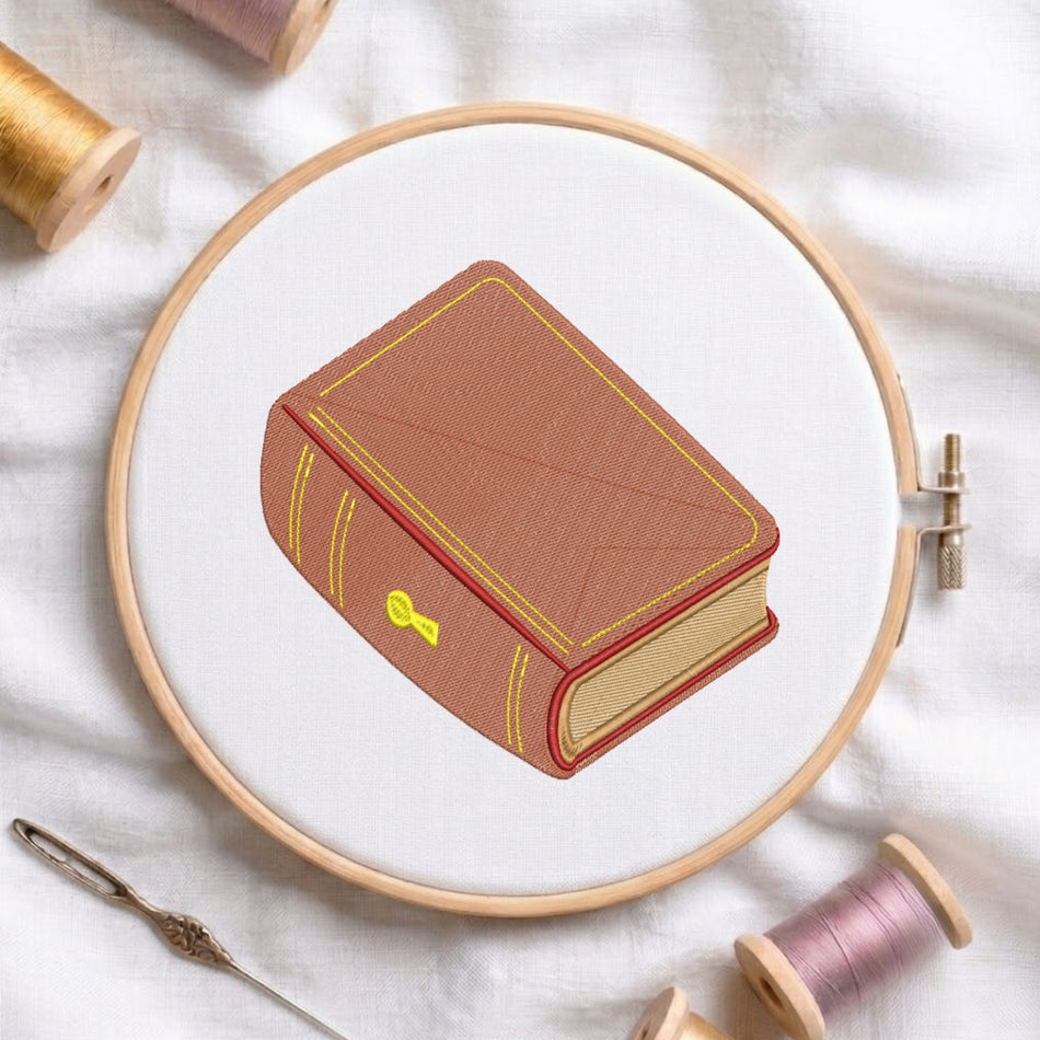 A circular embroidery hoop on white fabric holds a stitched illustration of a brown book with yellow trim and a small yellow key detail. Spools of thread and a needle are arranged around it.